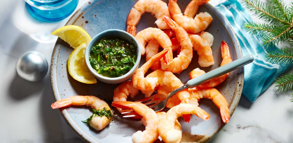 Pile of shrimp scattered on a blue plate next to a bowl of gremolata.
