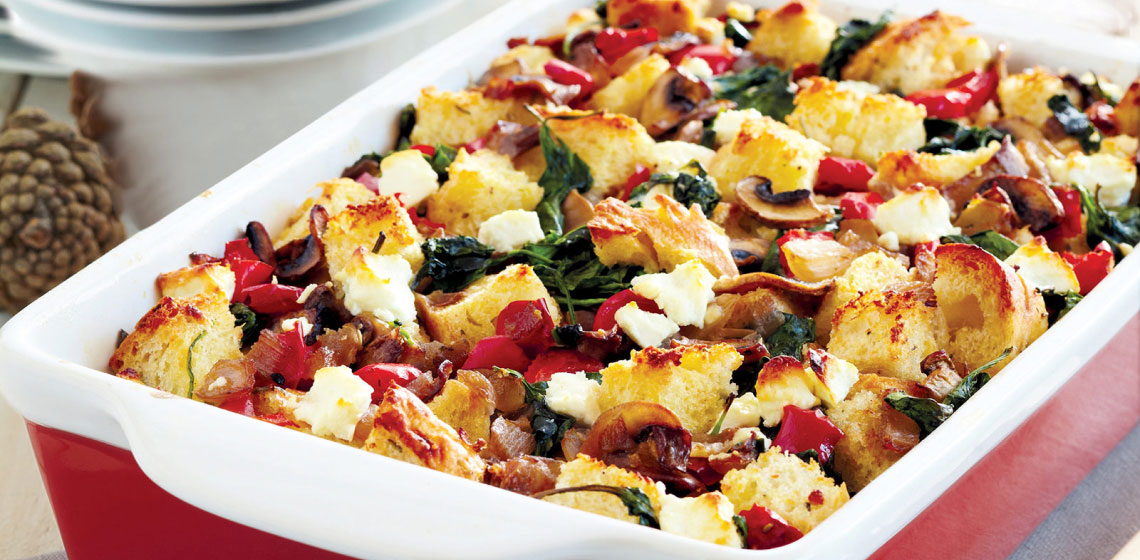 A white rectangular baking dish full of a mushroom, spinach, and goat cheese strata dotted with torn pieces of bread and red peppers.