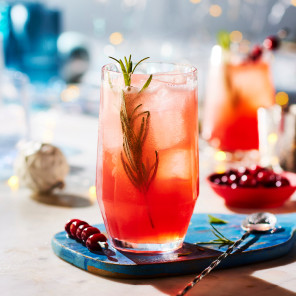 A highball glass filled with cranberry pink cocktail, garnished with a rosemary sprig, on a white marble surface scattered with holiday decorations.