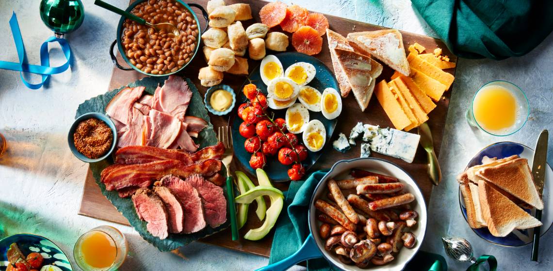 A big wooden charcuterie board loaded with soft-cooked eggs, sausages, bacon, bread, cheese, pickles, and a green bowl of baked beans.