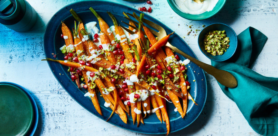Oval blue platter of roasted carrots with yogurt tahini sauce, chopped pistachios, and fresh pomegranate arils