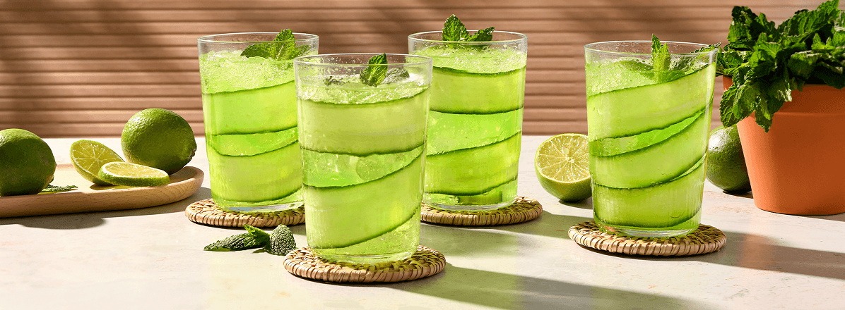 Four lime rickey sodas with a cucumber ribbon lining the glass and a sprig of mint on the top. Side of sliced limes on a wooden board.