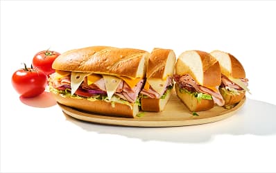 Dagwood sandwich filled with assorted deli meats, cheese, and veggies on a wooden cutting board. Half of the sandwich is cut into slices, showing the cross section. 
