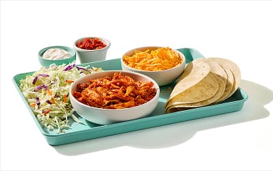 Tray holding a bowl of al pastor chicken, a bowl of shredded cheese, folded tortillas, and a bed of shredded cabbage. On the side, two small dipping bowls of sour cream and salsa. 