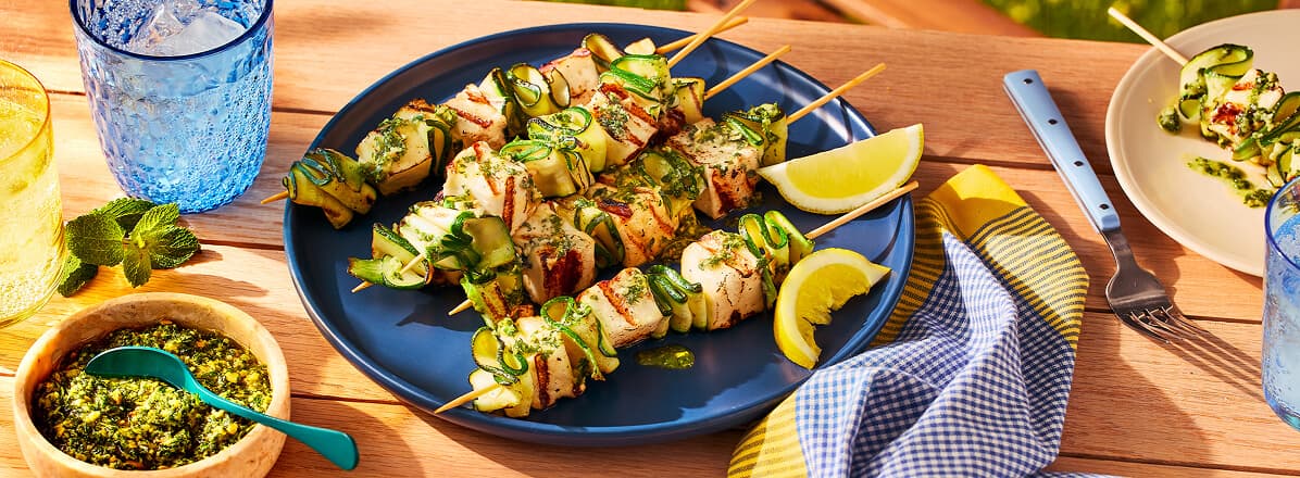Grilled zucchini ribbon and halloumi skewers with a lemon mint pesto drizzle on a blue plate with lime wedges on the side.
