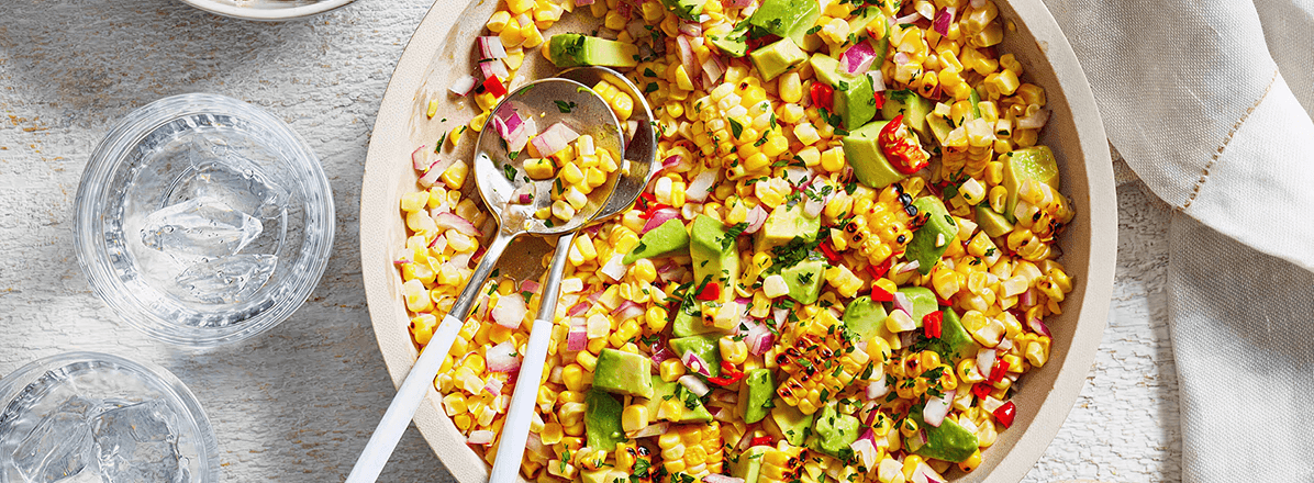 Overhead shot of Avocado & Corn Salad in a white serving bowl with two glasses of water and a small plate of cut limes next to it.