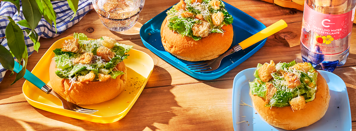 Three caesar salads in bread bowls on colourful plates on a picnic table