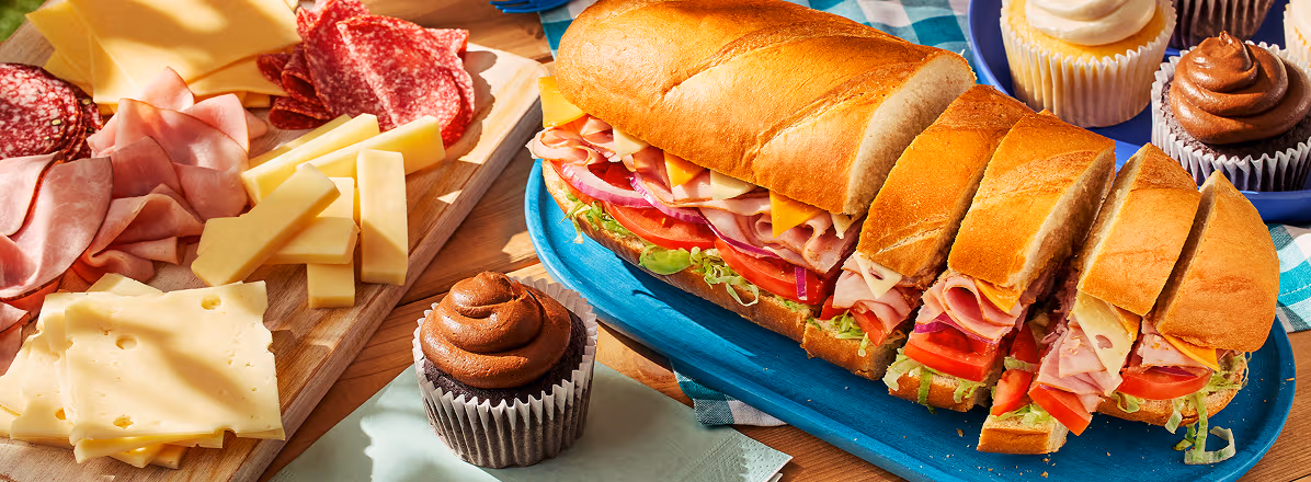 Classic picnic set up with Dagwood sandwich, and charcuterie board to the side. Chocolate cupcake on a plate in the foreground.