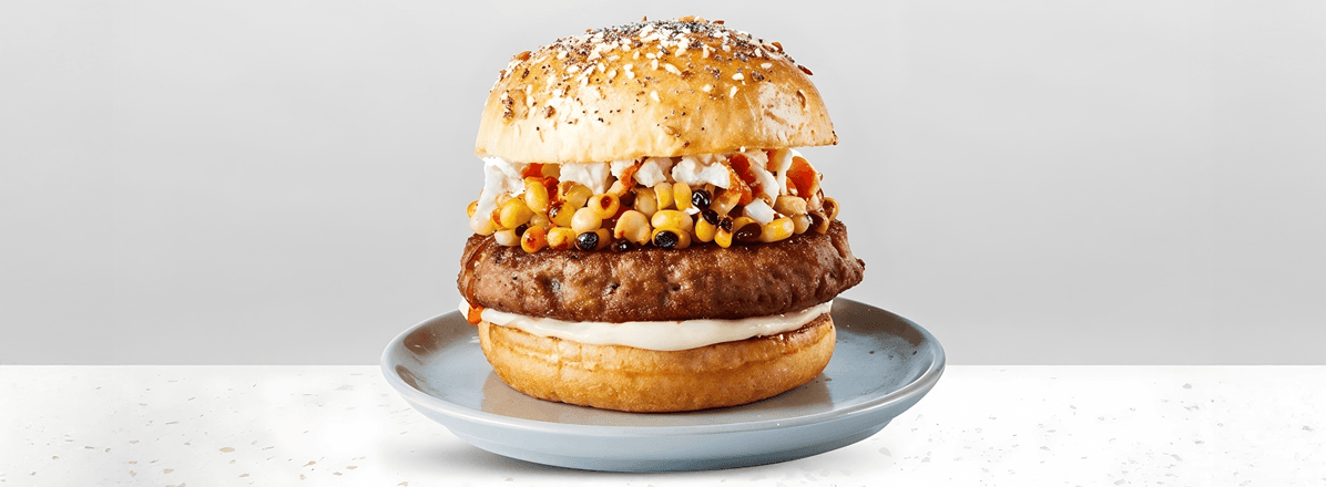 Seeded bun topped with a turkey burger patty, grilled corn, hot sauce, mayo, and crumbled feta cheese sitting on a blue gray plate.