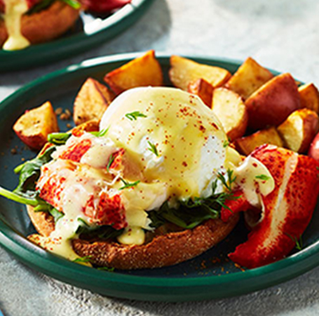 Eggs benedict with lobster, spinach and hollandaise sauce and side of crispy potatoes on green plate.