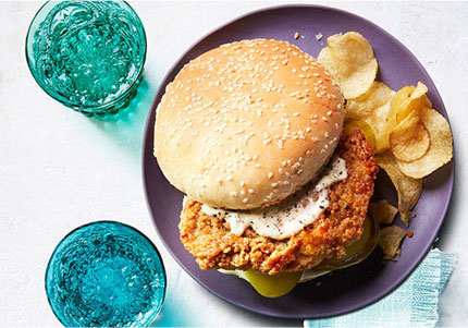 Sliced chicken sandwich on purple plates with side of potato chips.