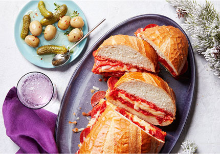 Sliced Dagwood sandwich on purple platter with side of pickles on small blue plate.