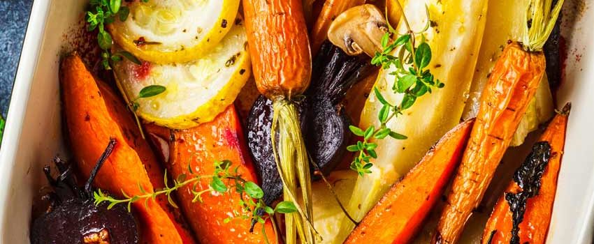 Various roasted root vegetables including carrots, sweet potatoes and beets in a white baking dish on a blue counter top.