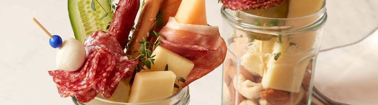 Two jars containing charcuterie meats and cheeses such as salami, cheddar, nuts and melon.