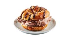 Close up of croissant filled with Coffee Crisp spread drizzled in fudge sauce with candy bar pieces on white plate.