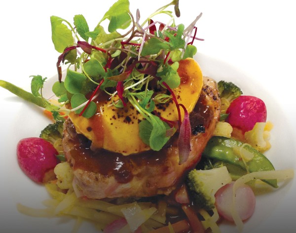 Nagano pork loin chop with port sauce, onion confit and goat cheese piled high on a white plate with sauce dripping down, topped with microgreens.