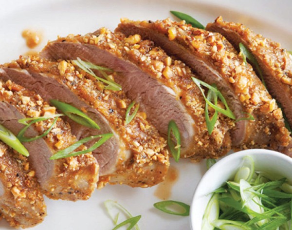 Peanut-crusted, sliced pork tenderloin atop a white serving platter, garnished with sliced green onions.