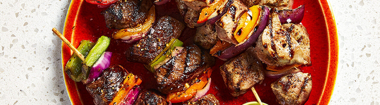 Grilled beef and pork kabobs on a red plate with a little ramekin of salsa verde.