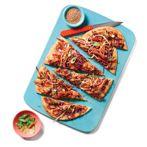 Korean-style, pulled pork topped flatbread garnished with coleslaw, green onions and sesame seeds sitting on a blue cutting board with a knife next to it.