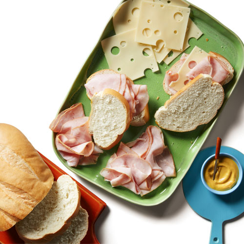 Black Forest ham, baguette bread slices, and sliced Swiss cheese on a green serving platter next to a little pot of yellow mustard with a spoon in it.
