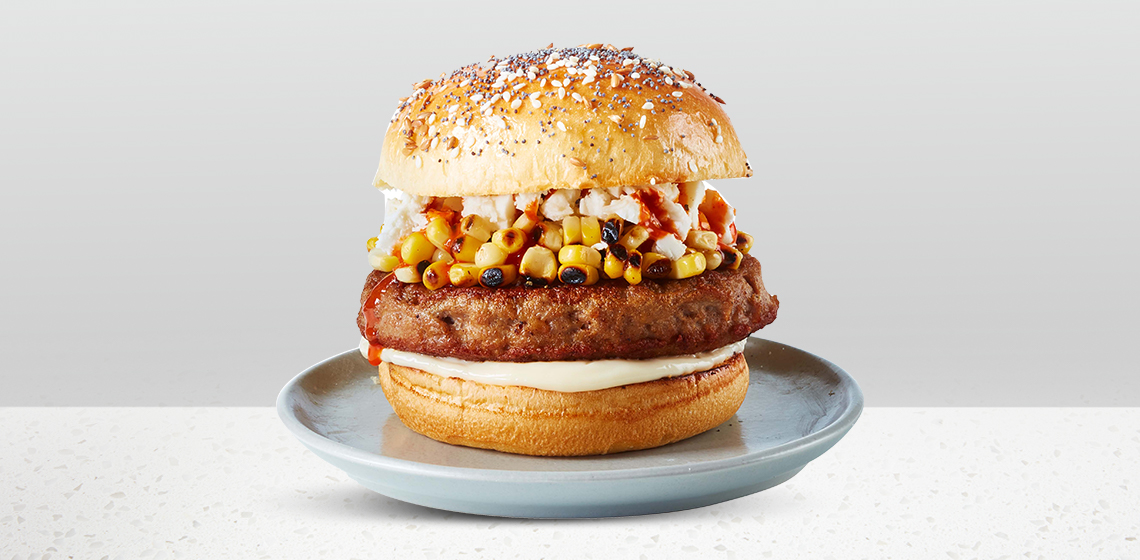 Seeded bun topped with a turkey burger patty, grilled corn, hot sauce, mayo, and crumbled feta cheese sitting on a blue gray plate.
