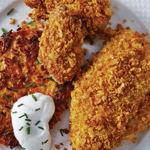 Cornflake crumb coated oven fried chicken on a white plate sitting next to a sweet potato and carrot rosti. 