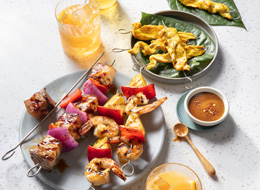 Three Pan-Asian style skewers including chicken, pork belly and shrimp on green-lined platter and a gray platter with orange beverage to one side. 