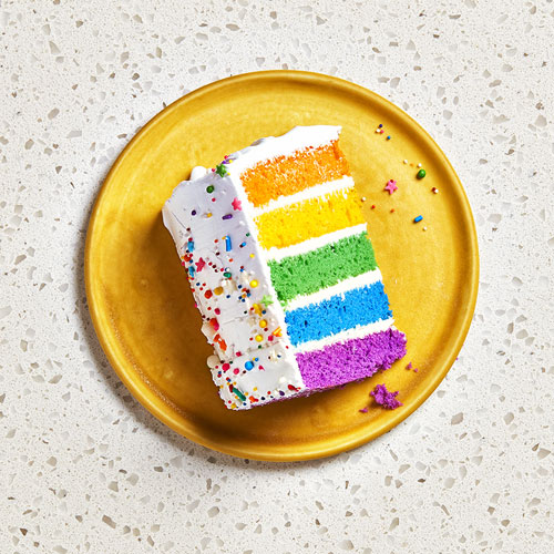  Plated shot of the Bakery Department's five-layer rainbow cake, iced in white buttercream with sprinkles, sitting on a yellow plate. 