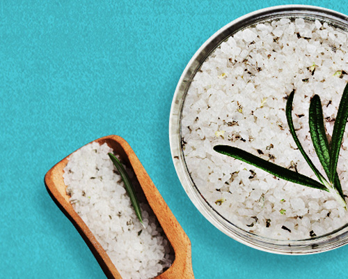 Silver bowl with rosemary-flecked salt and a wooden scoop next to it filled with the same herbed salt.