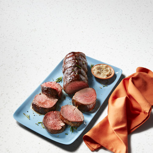 Grilled Sterling Silver beef medallions sliced on a blue plate with a little bowl of horseradish sauce next to it.