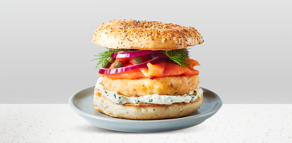 An everything spiced bagel topped with a fresh sockeye salmon burger patty, smoked salmon sliced, herbed cream cheese, red onion slices, capers and dill sitting on a blue gray plate.