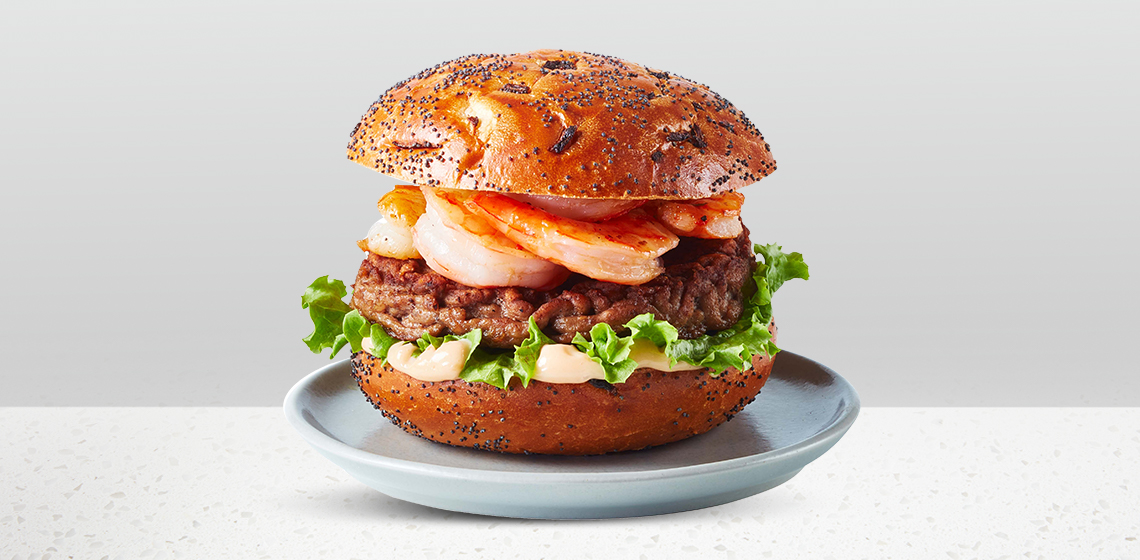 Onion bun topped with a sirloin beef burger patty, grilled shrimp, seafood sauce, mayonnaise and greens sitting on a blue gray plate.