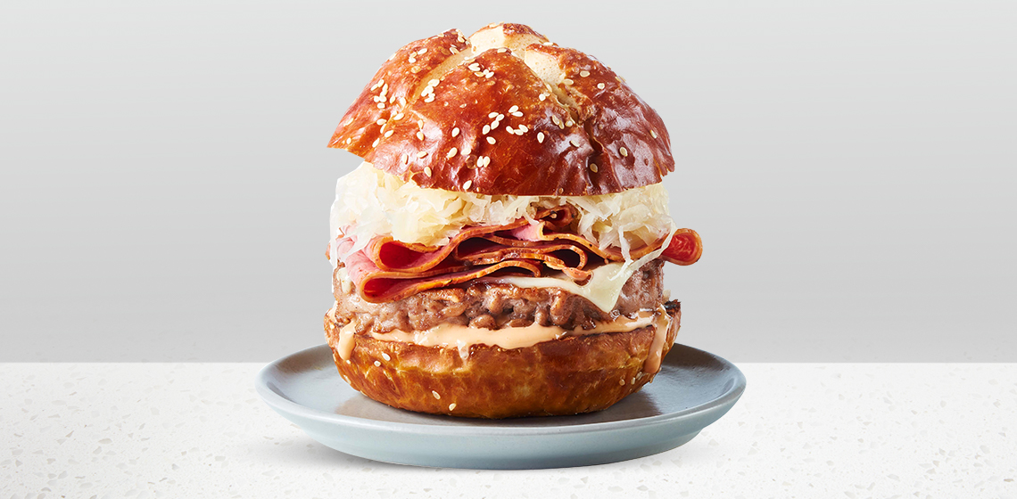 Pretzel bun topped with a Compliments prime rib burger, sauerkraut, Compliments Thousand Island Dressing, Swiss cheese and deli pastrami slices, sitting atop a blue gray plate.