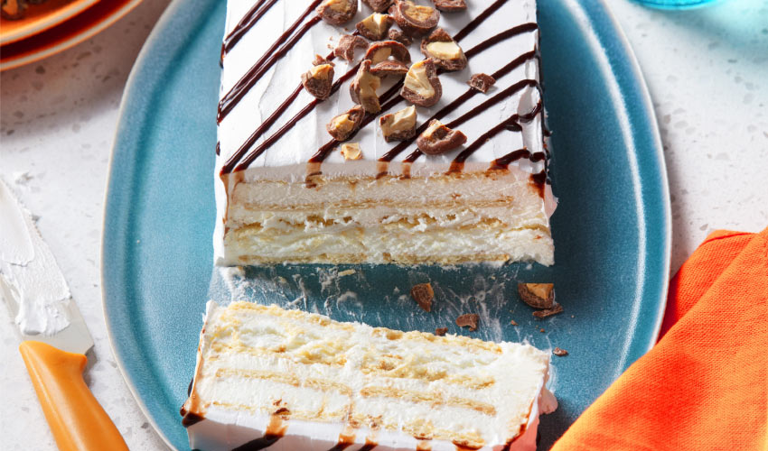Ultimate-Choco-Caramel-Ice-Cream-Sandwich-Cake Choco-caramel layered ice cream cake using Compliments Caramel Flavour ice cream sandwiches, topped with a drizzle of sundae chocolate topping on a blue plate with the first slice cut.