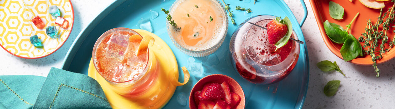 Top down view of our three of our zero proof mocktail recipes sitting on an orange and a blue tray with garnishes including fresh herbs and strawberries.