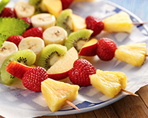Threaded pieces of fruit including strawberries, pineapple, and kiwi fruit on a blue and white plate. 