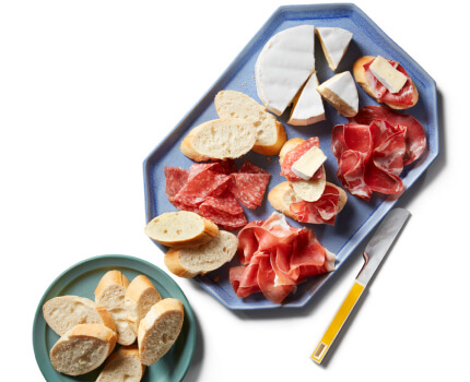 Italian cold cuts, baguette bread slices, and a wheel of Brie cheese on a blue serving platter next to a little green plate with bread on it. 