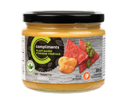 Clear glass jar of Compliments plant-based queso dip pictured was a dip on red tortilla chips on the jar. 