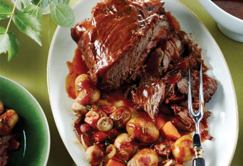 A multicooker prepared pot roast on a white platter surrounded by cooked potatoes and button mushrooms on a table scape. 