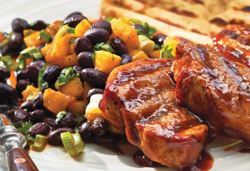 Smoky, sauce glazed pork medallions on a white plate sitting next to a black bean and pineapple salad.
