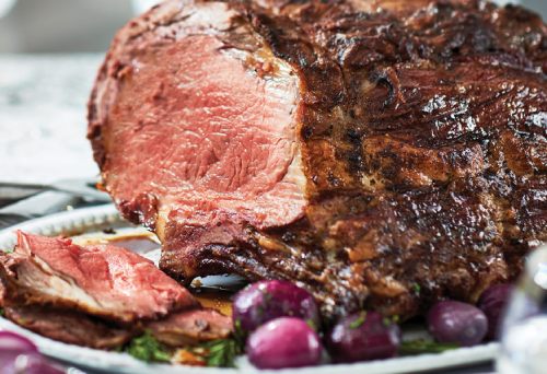 Whole salt-crusted Sterling Silver prime rib on a platter surrounded by roasted and fresh vegetables on a dressed table setting.