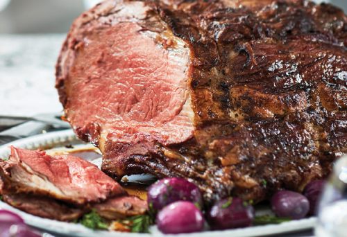 Whole salt-crusted Sterling Silver prime rib on a platter surrounded by roasted and fresh vegetables on a dressed table setting.
