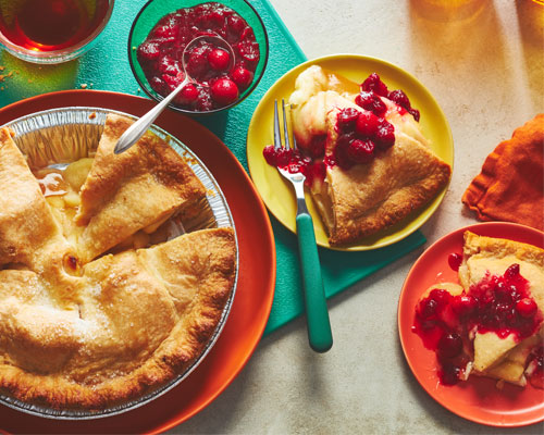 Baked apple pie sitting next to a slice with a spiced cranberry sauce drizzled on top.