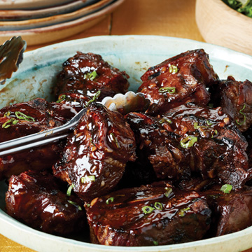  A light-blue bowl full of glazed Asian-style BBQ beef short ribs with tongs for serving.