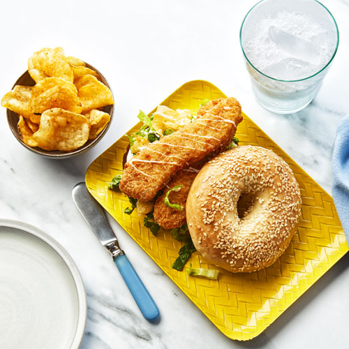 Chicken finger and pickle topped sesame bagel on a rectangular yellow plate.
