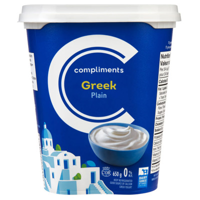 A white tub of Compliments Plain 0% Greek Yogurt with an illustration of a Greek village and whitewashed houses by the sea.