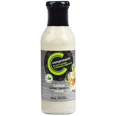 A bottle of Compliments Plant-Based Caesar Dressing with a black label and a wedge of dressed lettuce on the side.
