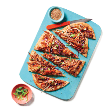  Korean-style pulled pork–topped flatbread garnished with coleslaw, green onions, and sesame seeds, sitting on a blue cutting board with a knife next to it.