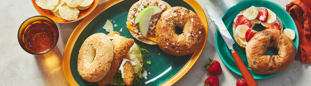 Three different bagels with different toppings including strawberries and banana, sit atop a blue and yellow plate with a side of chips.