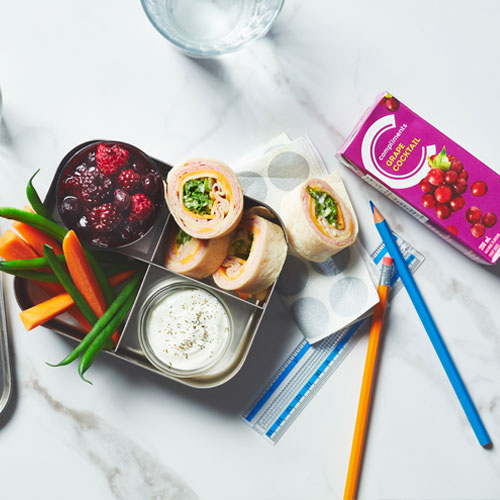 Metal bento box filled with ham and cheese wraps, veggies and dip and a field berry compote with a juice box on the side.